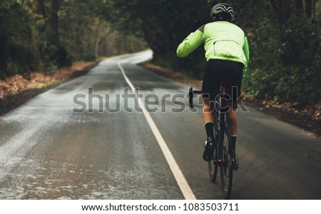 Rear view of male athlete cycling on country road on rainy day. Professional cyclist riding a bike on empty highway through forest.