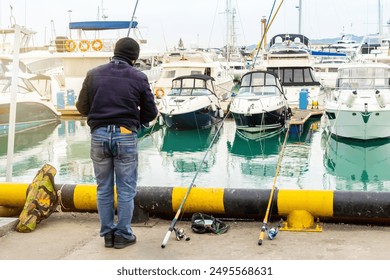 Rear view of a male fisherman using a fishing rod in the marina of the seaport. A marine berth for mooring ships. The ships are docked in the port. - Powered by Shutterstock