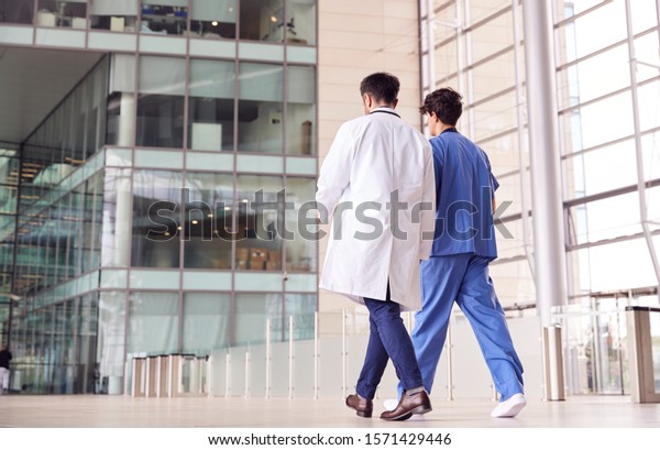 Rear View Of Male Medical Staff\
Talking As They Walk Through Lobby Of Modern Hospital\
Building