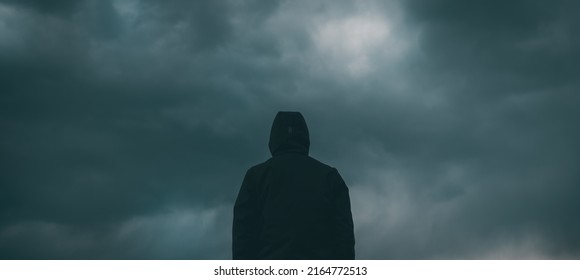 Rear view of male person wearing hooded jacket against dark moody dramatic clouds at sky, man looking into uncertain ominous future - Shutterstock ID 2164772513