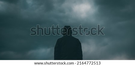 Rear view of male person wearing hooded jacket against dark moody dramatic clouds at sky, man looking into uncertain ominous future