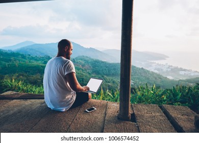 Rear View Of Male Traveler Blogger Work Remote On Netbook Computer While Enjoying Amazon Nature Landscape View Outdoors. Young Man Using Online Banking For Sending Money Everywhere You Are From Laptop