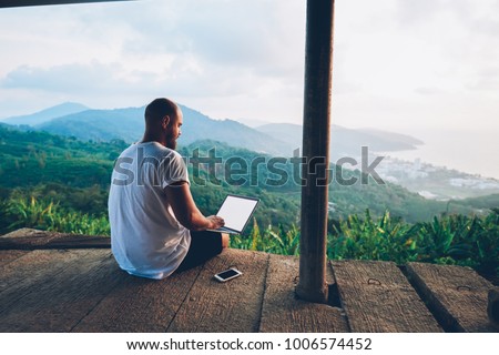 Rear view of male traveler blogger work remote on netbook computer while enjoying amazon nature landscape view outdoors. Young man using online banking for sending money everywhere you are from laptop