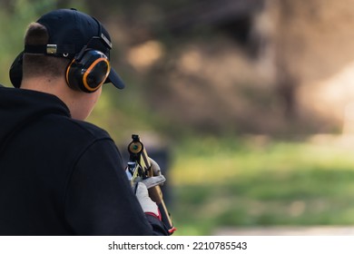 Rear View Of Man In Black Hoodie And Cap Wearing Safety Headphones Reloading Gun. Gun Training. Firing Range. Outdoor Horiozntal Shot. High Quality Photo