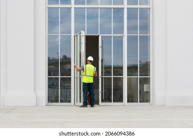 A Rear View Of A Man In A Construction Helmet And Reflective Vest Standing At The Glass Doors And Holding A Folding Ruler.