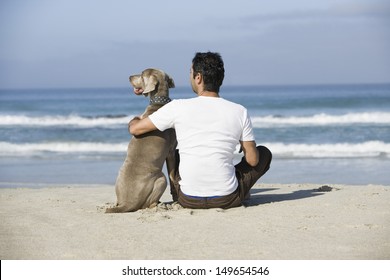 Rear View Of A Man And Dog Sitting On Beach