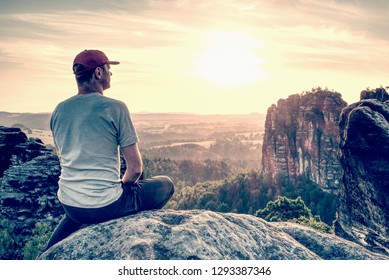 Rear view of man hiker sitting on rock on top of hill while looking at sunset over valley.