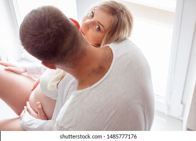 Rear view of the man hugging the belly of pregnant woman. People are wearing white clothes. Husbant kiss the wife