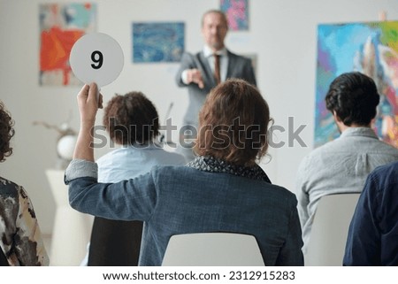 Rear view of man raising his tablet with number up while sitting at auction in art gallery among other people