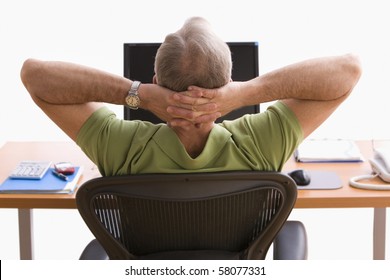 Rear View Of A Man Seated At A Desk In Front Of A Laptop. He Is Sitting Back In His Chair With His Hands Behind His Head. Horizontal Shot.