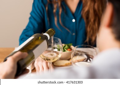 Rear View Of A Man Serving White Wine In A Glass. Young Couple Enjoying Meal And Drinks At Home. Close Up Of Man Hand Pouring Wine At His Girlfriend.