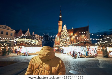 Rear view of man in warm clothing during night walk in city. Traditional Christmas market on Town Hall Square in Tallinn, Estonia.