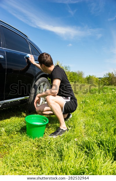 Rear View of Man Washing Car with Soapy Sponge,\
Crouching Next to Green Bucket in Green Grassy Field on Bright\
Sunny Day with Blue Sky