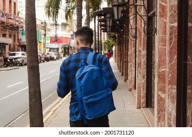 Rear view of a man wearing a backpack and blue plaid shirt walking through the city streets.
