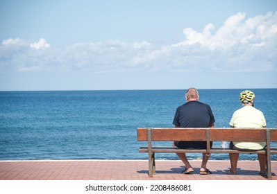 Rear View Of A Mature Couple Sitting On A Bench Overlooking The Sea