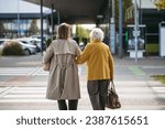 Rear view of mature granddaughter walking arm in arm from shop. Caregiver carrying groceries to senior woman