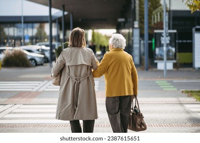 Rear view of mature granddaughter walking arm in arm from shop. Caregiver carrying groceries to senior woman's car. Elderly lady shopping at the shopping center, needing help loading groceries into