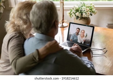 Rear view mature grey haired couple man and woman making video call to relatives, using laptop, hugging, sitting at table, grown up children communicating with elderly parents, virtual communication