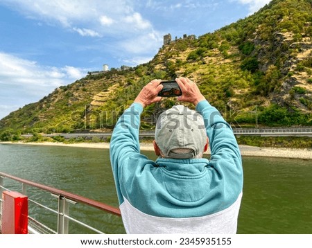 Rear view of a mature man using his mobile phone to capture a picture of the beautiful landscape and medieval castle. He is standing on the deck of a boat cruising on the Rhine River.