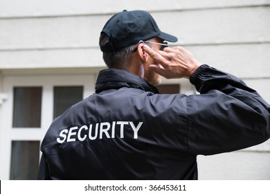 Rear View Of Mature Security Guard Listening To Earpiece Against Building