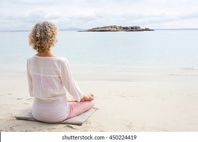 Rear view of mature woman in holiday destination beach contemplating blue sea sitting on shore in yoga position, meditating outdoors. Well being mature sport and healthy lifestyle, nature exterior. - Powered by Shutterstock