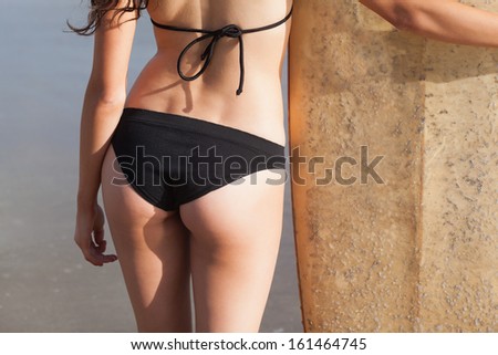 Rear view mid section of a slender woman in black bikini bottom with surfboard on beach