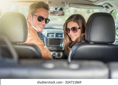 Rear View Of A Middle Aged Couple In A Black Seats Car. They Are Looking At Camera Wearing Their Leather Coats And Sunglasses. The Grey Hair Man With Beard Is In The Driver Seat