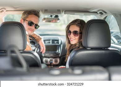 Rear View Of A Middle Aged Couple In A Black Seats Car. They Are Looking At Camera Wearing Their Leather Coats And Sunglasses. The Grey Hair Man With Beard Is In The Driver Seat