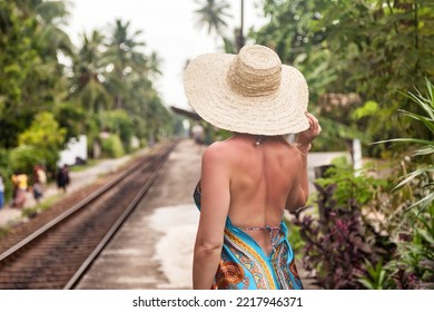 Rear View Of Middle Aged Woman Standing On Railway Station Waiting Train. Female In Summer Dress And Sun Hat In Tropical Background With Palm Trees. Travel Vacation Concept. Copy Text Space