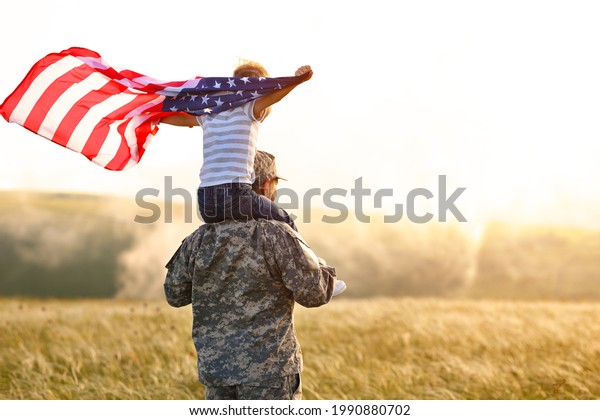 Rear view of military man father carrying happy\
little son with american flag on shoulders and enjoying amazing\
summer nature view on sunny day, happy male soldier dad reunited\
with son after US army