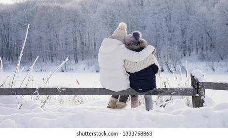 Rear View Mom And Daughter In Warm Clothes Hugging Each Other Admiring The Winter Landscape In A Mountainous Area Sitting On A Snow-covered Wooden Fence.