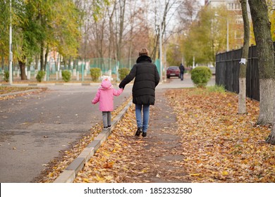 Rear View Of Mother And Her Little Daughter Walking Along City Street. Woman Leading Child By Handle Along Curb In Autumn Season