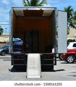 Rear View Of A Moving Truck With Back Doors Open And Loading Ramp Down, Showing Storage Containers And Moving Blankets Inside.
