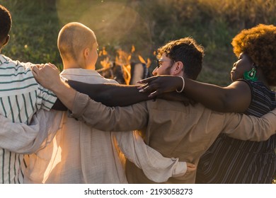 Rear View Of Multicultural Good Old Friends Hugging And Having A Weekend Picnic In Nature Near The Campfire, Talking And Laughing. Sunset Time. High Quality Photo