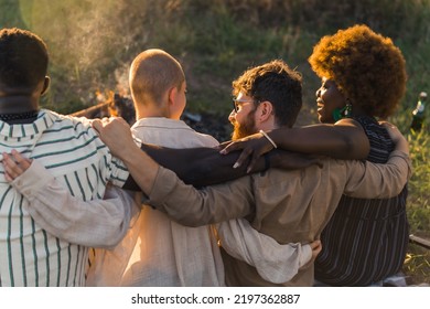 Rear View Of Multicultural Good Old Friends Hugging And Having A Weekend Picnic In Nature Near The Campfire, Talking And Laughing. Sunset Time. High Quality Photo