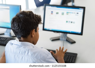 Rear view of multiethnic boy using desktop computer in school library. Back view of mixed race child working in classroom with computer at elementary school. Young arab schoolboy typing on keyboard.