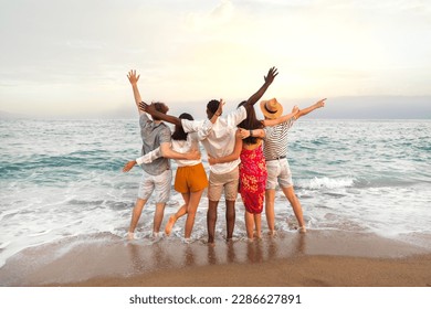 Rear view of multiracial friends embracing together looking at the ocean celebrating with arms up during vacation trip. - Powered by Shutterstock