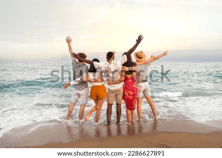Rear view of multiracial friends embracing together looking at the ocean celebrating with arms up during vacation trip.