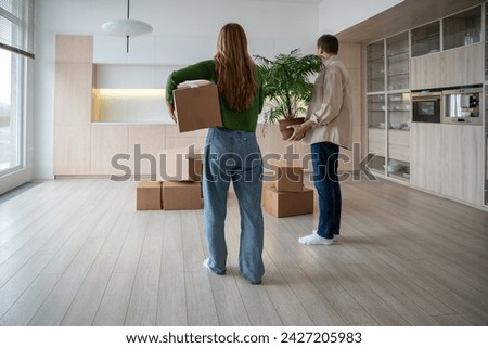 Rear view of new home owners with boxes and houseplant in hands. Family couple taking things to another rented light apartment. Housewarming in mortgage apartment. Beginning of happy married life.