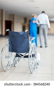 Rear View Of Nurse Helping Patient Walk With Wheelchair In Foreground 