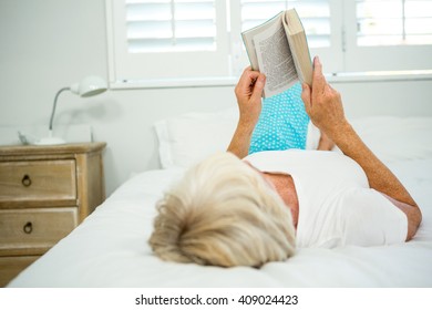 Rear View Of Old Woman Holding Book On Bed Against Window At Home