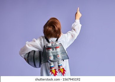 Rear view on child wearing protective suit holding helmet and showing up at cosmos, at stars. Portrait, isolated over purple background