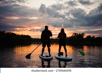 rear view on couple of people standing on sup boards with oars in their hands on the river at sunset