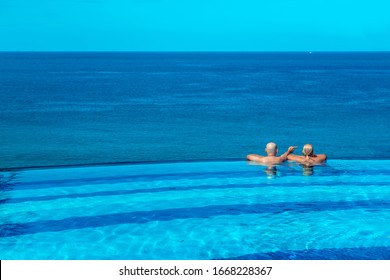 Rear View On The Edge Of The Infinity Pool: Couple Admiring The Sea View. A Man Points To A Beautiful Boat In The Sea