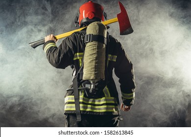 Rear View On Fire Fighter Stand Holding Hammer Isolated Over Smoky Background, Wearing Red Helmet