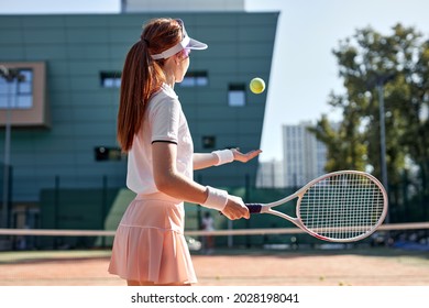 Rear view on forceful woman playing tennis in outdoor court, hitting ball, sport concept. Slender redhead lady in short skirt and cap is concentrated on game during competition, view from back
