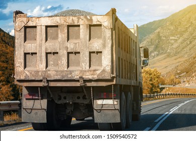 Rear View On A Large Dump Truck Rides On A Highway In The Mountains While Transporting Goods Over Long Distances. Fast Delivery By Ground Transportation.