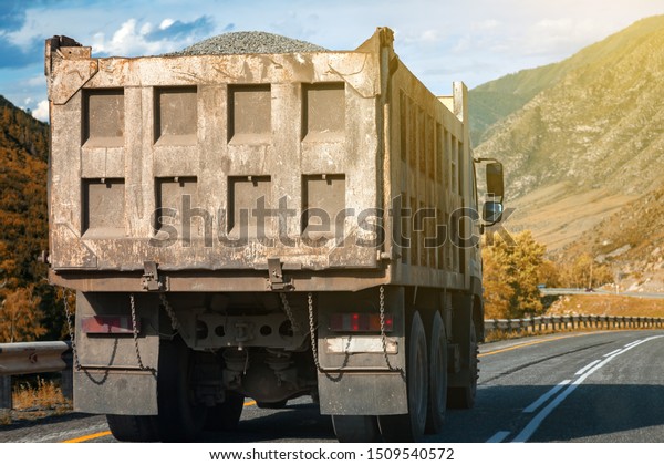 Rear view on a large dump truck\
rides on a highway in the mountains while transporting goods over\
long distances. Fast delivery by ground\
transportation.