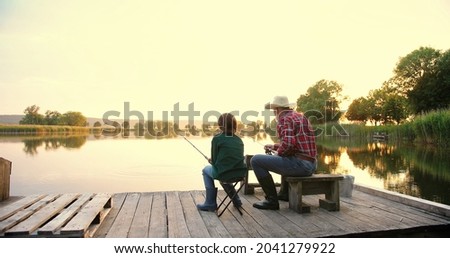 Rear view on the old grandfather and his small grandson fishing together and throwing rods in the lake while sitting on the wooden bridge or pier at the sunset