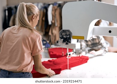 Rear view on young caucasian blonde seamstress working on sewing machine in textile factory alone. Female controlling automated production of clothing in fabric. design, tailoring industry - Powered by Shutterstock